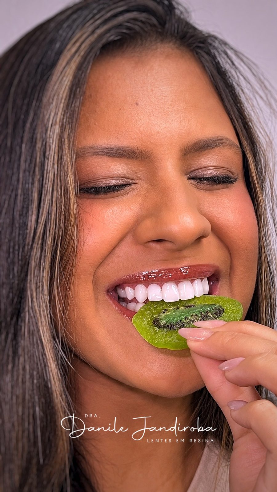 Mulher sorrindo de olhos fechados ao morder uma fatia de kiwi, mostrando os dentes da frente